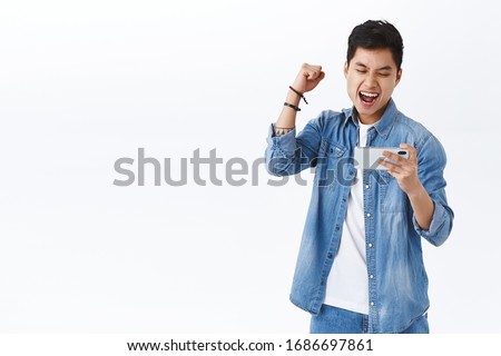 Technology, online lifestyle concept. Triumphing asian rejoicing man winning in game, raising fist up in celebration saying yes, hooray, smiling pleased at mobile phone, finished level first