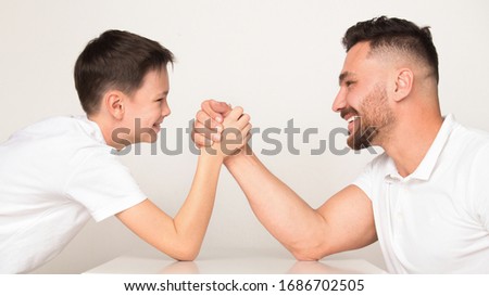 Playful dad and son compete in arm wrestling, light studio background, panorama