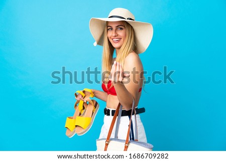 Young Uruguayan blonde woman in swimsuit in summer holidays holding a beach bag and summer sandals over isolated blue background inviting to come with hand. Happy that you came