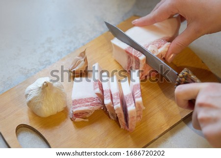 Horizontal indoor picture of unknown person holding knife, cutting lard into slices, preparing for lunch. Salo and garlic being on wooden cutting board, traditional food. Eating habits concept.