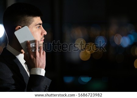 Serious Businessman Talking On Mobile Phone Standing Near Panoramic Window In Modern Office At Night. Side View, Copy Space