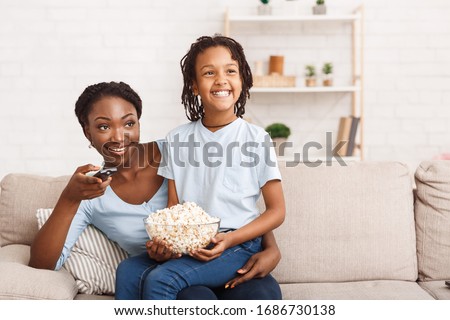 Family Time Concept. Happy african mom and child sitting on sofa, eating popcorn and watching tv, empty space