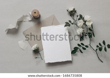 Moody wedding styled composition. Feminine desktop mockup scene with white rose flowers and leaves, silk ribbon, craft envelope and blank greeting card on grey textured background. Flat lay, top view.