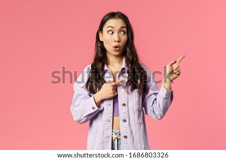 Portrait of enthusiastic, excited pretty asian woman in denim jacket, open mouth in surprise, say wow seeing cool thing, pointing and looking right, standing pink background astounded