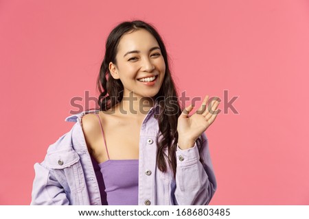 Close-up portrait of happy good-looking asian girl saying hi, waving hand casually, informal greeting sign, laughing and smiling friendly, welcome friend, greet hello, stand pink background