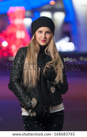 Fashionable lady wearing cap and black jacket outdoor in xmas scenery with blue lights in background. Portrait of young beautiful woman with long fair hair posing smiling in winter style.