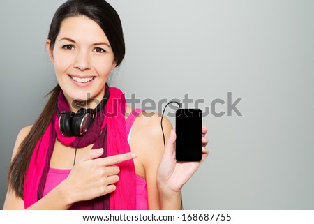 Portrait of an attractive happy brunette young woman wearing headphones around her neck and pointing to her smart mobile phone, on grey background