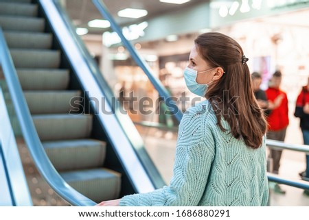 beautiful young woman rides the escalator of a shopping center with a protective mask on her face from the virus -infected air. The concept of protection from coronavirus while shopping. Royalty-Free Stock Photo #1686880291