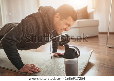 Young ordinary man go in for sport at home. Wink and scream because of hard exercises for freshman or ordinary guy at beginning. Try to do push ups or stand in plank position Royalty-Free Stock Photo #1686917254