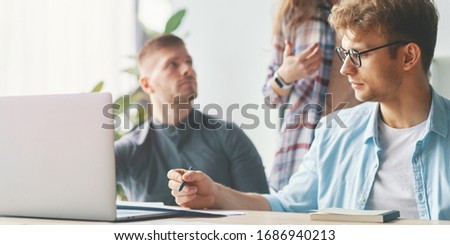 Side view of concentrated at work confident young man in smart casual wear working on laptop while sitting near window in creative office with other workers on background