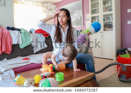 Stressed mother working from home with messy room and her little baby playing with many toys. Asian housewife tired and suffering from headache overwork. Royalty-Free Stock Photo #1686972187