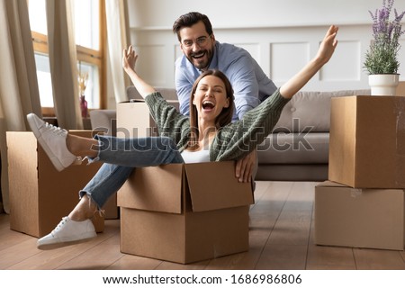 Full length overjoyed young bearded man in glasses pushing laughing wife in carton box. Energetic happy woman sitting in carboard container, having fun with smiling husband in new apartment house. Royalty-Free Stock Photo #1686986806