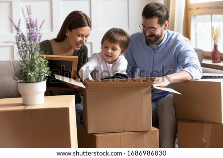 Smiling small child boy unpacking things together with affectionate parents in new living room. Happy young family couple helping little son unboxing belongings after moving in new apartment flat.