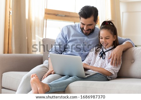 Smiling young father cuddling little schoolgirl daughter, watching funny movie together in living room. Happy small kid girl resting on couch with affectionate daddy, enjoying leisure time at home.