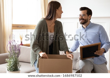 Overjoyed young married couple unpacking belongings from cardboard boxes, moving into new apartment flat. Happy 30s woman unboxing things, decoration items with smiling bearded husband in glasses.