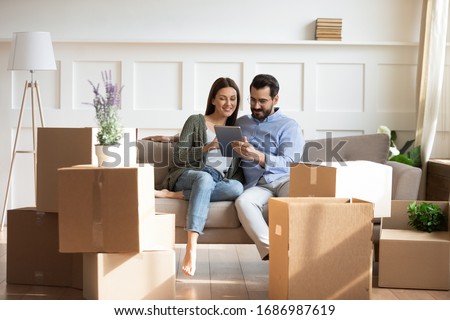 Full length happy family sitting on couch among cardboard boxes in new house, using digital tablet, buying decorations online. Smiling couple enjoying moving in new apartment, shopping in internet. Royalty-Free Stock Photo #1686987619