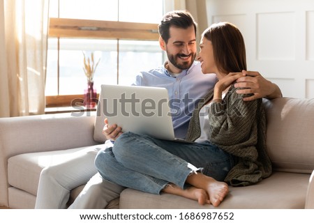 Smiling young bearded husband cuddling affectionate wife, watching romantic movie on computer at home. Loving family couple spending weekend free leisure time together, using laptop in living room.