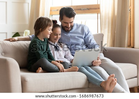Smiling young dad resting on couch with cute children, showing educational applications on computer. Happy man enjoying spending free leisure time with kids son daughter, watching movie at home.