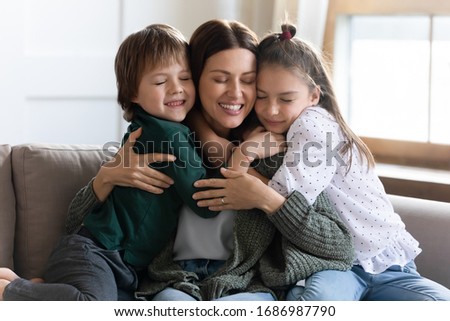 Affectionate small children embracing young mother, missing her after long separation. Loving mommy nanny babysitter cuddling little kids, relaxing together on comfortable couch in living room. Royalty-Free Stock Photo #1686987790