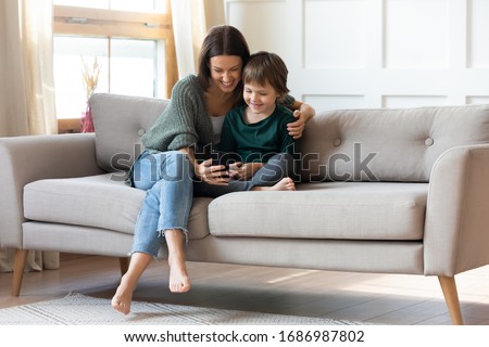 Full length smiling young barefoot woman hugging small cute child, taking selfie photo together on smartphone. Pleasant happy mommy resting on sofa with little kid boy, showing funny mobile apps.