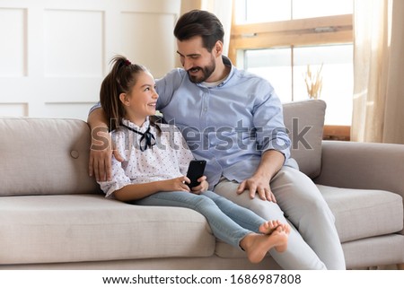 Smiling daddy cuddling pretty cute school aged daughter, using mobile phone together. Happy kid girl showing funny video to affectionate young father, resting on comfortable sofa in living room.