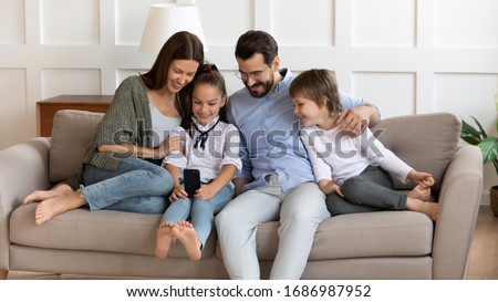 Front view joyful little child girl holding cellphone, showing funny video to laughing parents and brother, resting on sofa. Happy couple with children using funny mobile apps, enjoy weekend time.