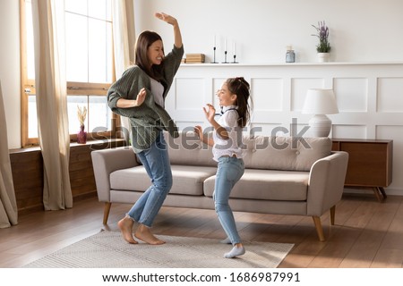 Full length overjoyed barefoot young mommy babysitter dancing to energetic music with school aged girl on floor carpet at home. Excited mother having fun with small child daughter in living room.