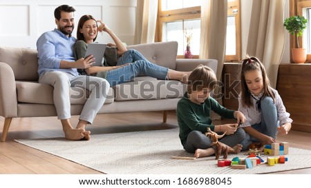 Full length overjoyed young couple resting on comfortable sofa, watching comedian movie on digital tablet, while little kids brother sister playing toys wooden blocks on floor carper in living room. Royalty-Free Stock Photo #1686988045