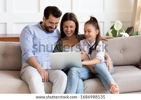 Happy bearded man embracing smiling wife and adorable school aged girl, watching funny video on computer. Affectionate family of three playing game on laptop or shopping online in internet store.