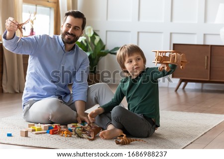 Playful young father holding dino, having fun with small kid son on floor in living room. Involved in interesting game little child boy playing with wooden airplane, enjoying playtime with daddy. Royalty-Free Stock Photo #1686988237