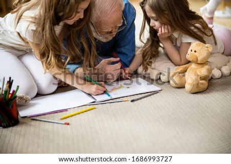 Twin girls entertaining grandfather by drawing with him at home on the carpet. Domestic concept