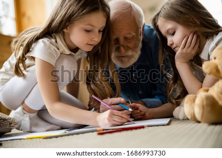 Pensive twin girls sitting comfortably on the carpet and drawing in a sketch book with their grandfather