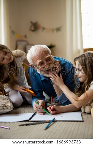 Happy grandpa laughing and grandchildren touching his beard while drawing with him. Domestic concept