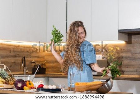 Happy young woman with eyes closed holding dill and parsley and dacing on the kitchen. Young housewife cooking. Healthy eating, vegetarian food, dieting and people concept. for weight loss and detox. Royalty-Free Stock Photo #1686994498