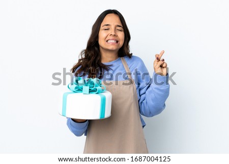 Pastry chef holding a big cake over isolated white background with fingers crossing and wishing the best