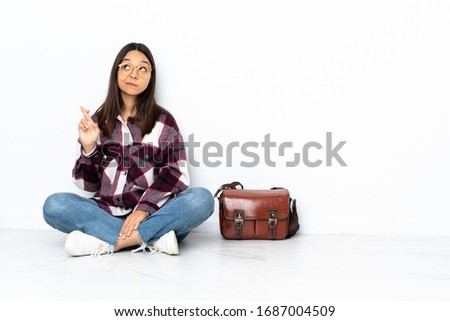 Young student woman sitting on the floor with fingers crossing and wishing the best