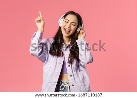 Portrait of attractive carefree asian girl chilling and vibing, listening music in headphones, raising finger up while trying sing along and reach highest note in song, pink background Royalty-Free Stock Photo #1687110187