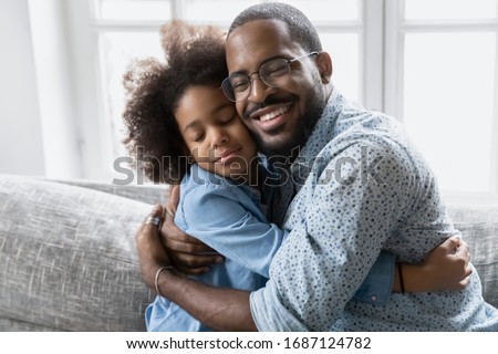 Loving African father in glasses hugs little daughter, family sitting on couch cuddling closed eyes enjoy sincere moment of tenderness, feeling bond express appreciation and gratitude for love concept Royalty-Free Stock Photo #1687124782