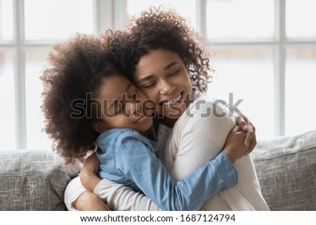 Seated on sofa african loving mother strong cuddles little daughter gives her love and caress show sincere candid feelings, concept of adopted kid and new caring mom, dear special person and affection Royalty-Free Stock Photo #1687124794