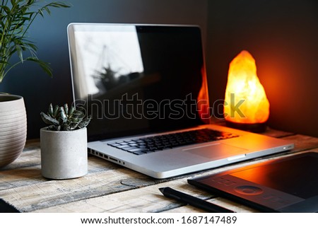 Cozy workspace with laptop, graphic tablet, plants and yellow glowing lamp at stone grey wall background. Working from home concept.