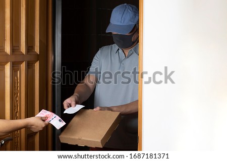 Pizza delivery man in blue baseball cap with mask on his face stands at door holding out pizza box and receipt, the customer's hand holds out a note of 100 Hong Kong dollars.