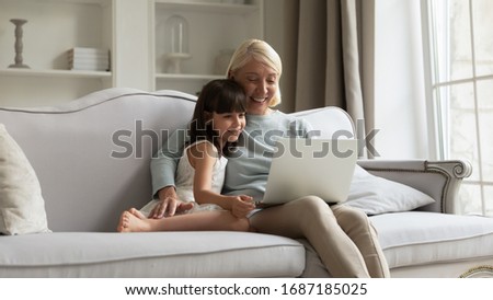 Happy elderly grandmother relax on couch in living room with little granddaughter watch video on laptop together, loving mature grandparent rest on sofa at home with small grandchild using computer