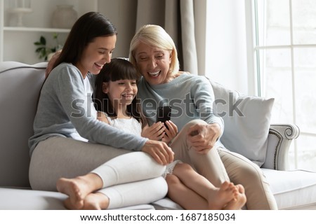 Overjoyed three generations of women sit rest on couch have fun watch funny video on smartphone together, happy little girl with grandmother and young mom relax on sofa at home using modern cellphone