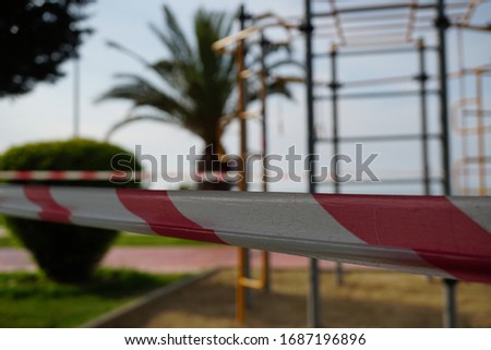   Barrier red and white tape closing a passage to a street sports ground, shot close-up on a background of sports equipment. COVID-19 pandemic. Palms, green vegetation, bicycle path on the seaside bou