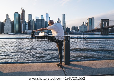 Athletic strong male in sportswear stretching leg and preparing for morning jogging while listening to music in earphones near river on urban background with skyscrapers and bridge