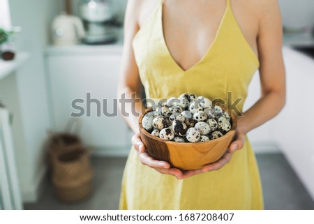 Woman in yellow dress holding in hands wooden bowl with pile of organic quail eggs. Easter cooking concept. Kitchen on background.