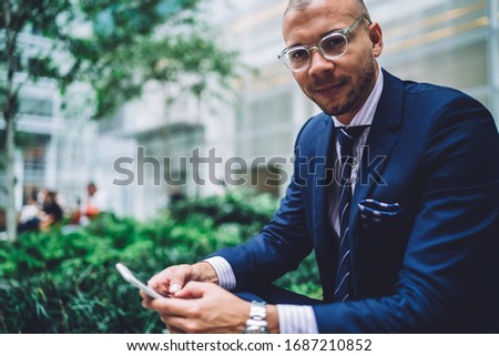Low angle of crop confident executive man in glasses and elegant suit looking at camera while sitting and interacting with smartphone on street against blurred exterior of modern office building