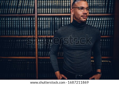 Content stylish student in gray turtleneck and glasses with hands in pocket looking away with smile nearby bookshelves in library