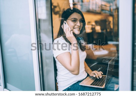 Side view through glass of cheerful smart young woman in glasses talking on cellphone while sitting with laptop on windowsill in college cafe
