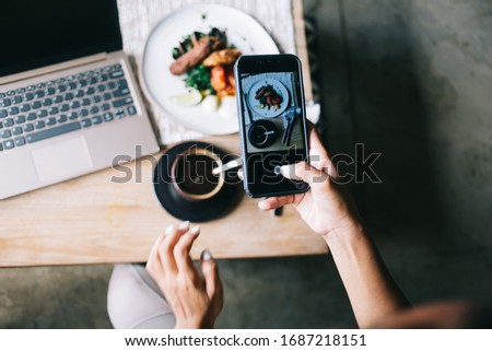 Crop from above female holding smartphone and taking photo of plate with food and cup of hot coffee next to opened silver laptop
